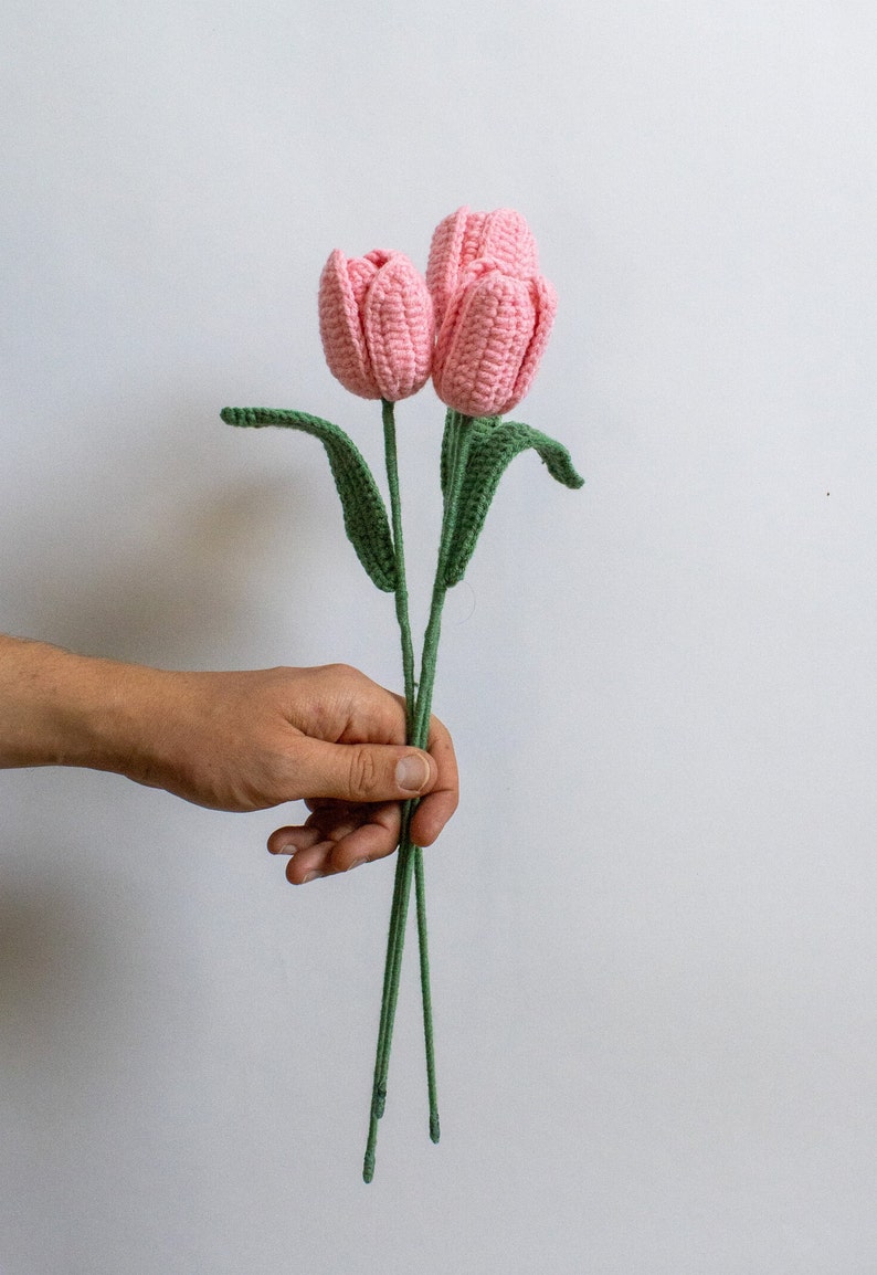 May include: Two pink crocheted tulips with green stems and leaves. The tulips are held in a hand.