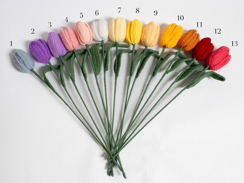 May include: A set of 13 crocheted tulips in various colors, including light blue, purple, pink, white, yellow, orange, red, and pink. The tulips are arranged in a fan shape with their stems bundled together.