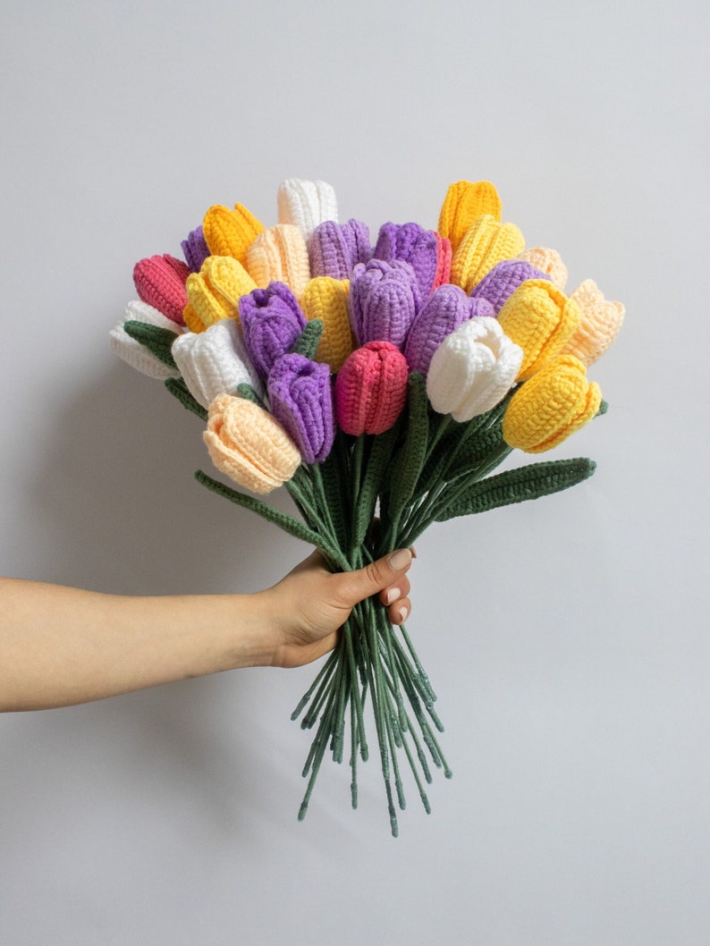 May include: A bouquet of crocheted tulips in shades of yellow, white, purple, and pink. The tulips are arranged in a hand-held bouquet with green stems.