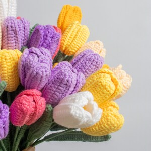 May include: A bouquet of crocheted tulips in shades of purple, pink, yellow, and white. The tulips are arranged in a hand-held bouquet with green stems.