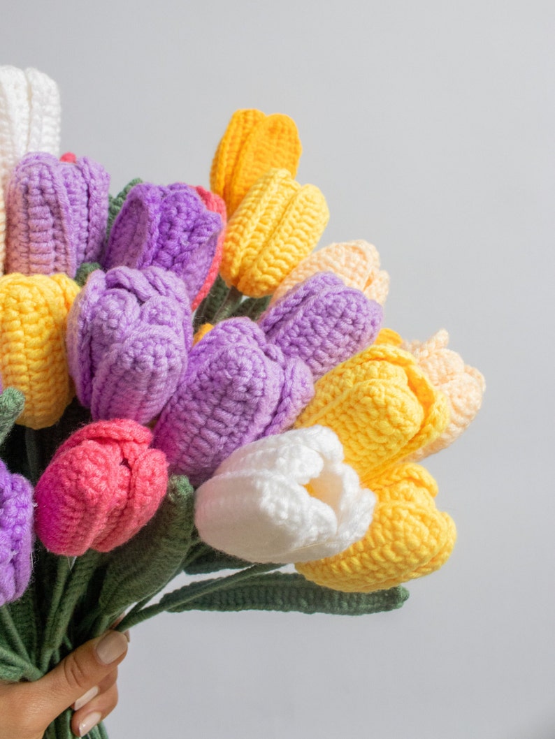 May include: A bouquet of crocheted tulips in shades of purple, pink, yellow, and white. The tulips are arranged in a hand-held bouquet with green stems.
