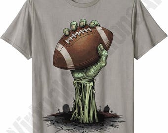May include: A light gray short-sleeve t-shirt featuring a graphic print of a green zombie hand emerging from cracked earth, holding a brown American football. The background of the graphic includes small tombstones in a graveyard setting.