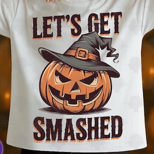 Puede incluir: Camiseta blanca con un diseño temático de Halloween. El diseño presenta una calabaza naranja con un sombrero de bruja negro, con las palabras "LET'S GET SMASHED" en letras marrones. La camiseta es de estilo casual y de manga corta.