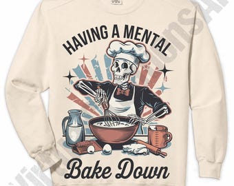 May include: A cream-coloured long-sleeve sweatshirt featuring a graphic of a skeleton wearing a chef hat and apron, whisking ingredients in a bowl. The text reads "HAVING A MENTAL BAKE DOWN" with illustrations of a pitcher, eggs, and a rolling pin.