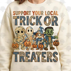 May include: Cream-colored sweatshirt featuring the text "SUPPORT YOUR LOCAL TRICK OR TREATERS" in orange and blue patchwork letters. The design includes illustrations of a mummy, ghost, witch, and Frankenstein's monster, each holding a jack-o'-lantern.