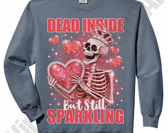 May include: A blue-grey sweatshirt with a graphic of a skeleton wearing a crown and holding a sparkling heart. The text "DEAD INSIDE" and "But Still SPARKLING" are in red. The design includes red hearts and stars.