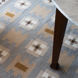 May include: A woven area rug featuring a repeating geometric pattern in shades of light blue, beige, white, and mustard yellow. The design consists of alternating square motifs and block shapes, finished with a simple linear border along the edge.