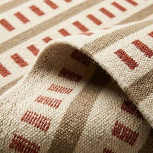 May include: A woven textile featuring a repeating pattern of horizontal stripes in tan and cream, accented by rows of small, rectangular terracotta-colored dashes. The fabric has a visible, coarse-textured weave with a folded edge showing depth.