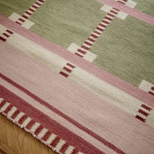 May include: A woven area rug featuring a geometric grid pattern in sage green, soft pink, and cream. The design includes horizontal and vertical bands with small, dark red stitched accents at the intersections and a fringed edge with red stripes.