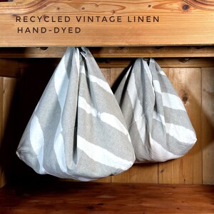 May include: Two hand-dyed, recycled vintage linen bags hanging on hooks. The bags are a light beige color with white stripes. The bags are hanging in a wooden cabinet.
