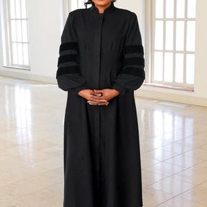 May include: A full-length image of a person wearing a black robe with a high collar and black velvet accents on the sleeves. The person is smiling and wearing black heels. The background is a bright room with arched windows.