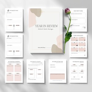 May include: A set of blank journal pages and a notebook titled "Year in Review" with a floral accent. The pages are designed for reflection and goal setting. A single purple flower with green leaves is placed next to the journal.