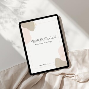 May include: A digital tablet showing a "Year in Review" template. The screen has text and abstract shapes in neutral tones. The tablet is on a beige fabric, with soft shadows. The text reads "Reflect. Reset. Realise."