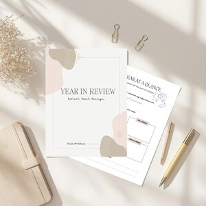 May include: A 'Year in Review' journal with the text 'Year in Review' and 'Reflect. Reset. Realign'. The journal is open to a page with a 'Year at a Glance' section. Two gold pens and paper clips are also visible.