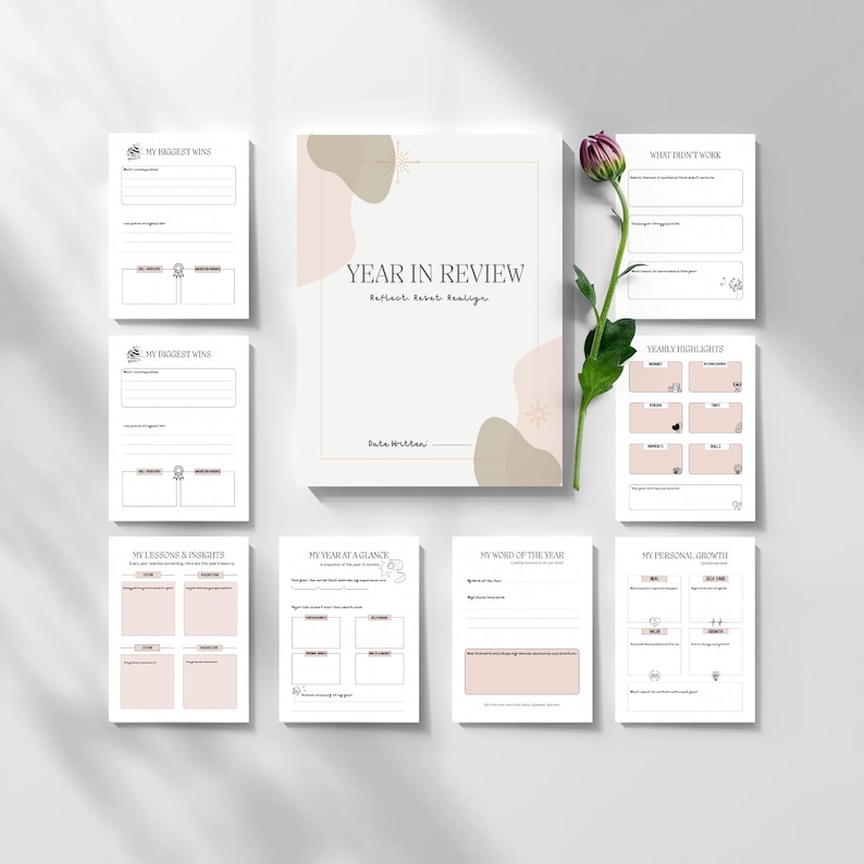 May include: A set of blank journal pages and a notebook titled "Year in Review" with a floral accent. The pages are designed for reflection and goal setting. A single purple flower with green leaves is placed next to the journal.