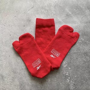May include: A pair of bright red split-toe socks featuring a white graphic print. The design includes the numbers 0 through 23 with the number 22 circled, the text Maison Margiela Paris, and a Reebok logo. The socks have a distinct tabi-style toe.