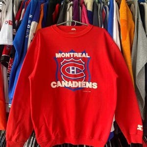Peut inclure: Un sweat-shirt rouge à manches longues et col rond arborant le logo de l'équipe de hockey des Canadiens de Montréal imprimé au centre. Le motif comprend le nom de l'équipe en blanc au-dessus et au-dessous d'un écusson bleu et rouge.
