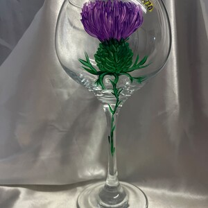 Peut inclure: Un verre à vin transparent orné d'un chardon violet peint à la main et d'une petite abeille. Le chardon a des feuilles et une tige vertes. Le verre est posé sur un fond blanc.