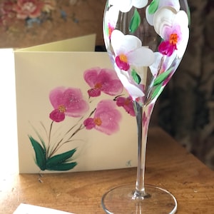 以下が含まれることがあります： ピンクと白の蘭が手描きされた、透明なガラス製のワイングラス。グラスは長いステムと丸いベースを持っています。