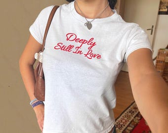 Könnte beinhalten: Weißes, bauchfreies T-Shirt mit dem Text "Deeply Still In Love" in roter, kursiv geschriebener Schrift. Das Model trägt eine silberne Halskette mit einem Herzanhänger.