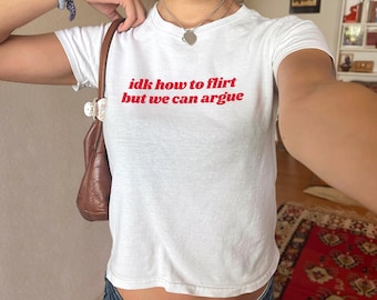 May include: White t-shirt with the red text "idk how to flirt but we can argue." The shirt is a crew neck style. The model is wearing a silver heart pendant necklace and a blue bracelet.