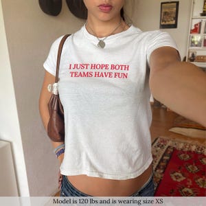 Op de afbeelding: Wit gecropped T-shirt met de tekst "I JUST HOPE BOTH TEAMS HAVE FUN" in rode letters. Het model draagt maat XS.
