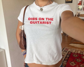 Könnte beinhalten: Weißes, bauchfreies T-Shirt mit dem Text "DIBS ON THE GUITARIST" in roten Buchstaben. Das Model ist 54 kg schwer und trägt Größe XS.