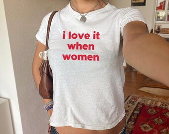 Könnte beinhalten: Weißes, bauchfreies T-Shirt mit dem Text "i love it when women" in roten Buchstaben.