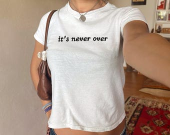 Könnte beinhalten: Weißes, bauchfreies T-Shirt mit dem Aufdruck "it's never over" in Schwarz. Das Model trägt eine silberne Halskette mit einem Herz-Anhänger.