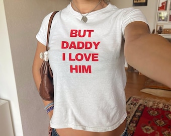 Puede incluir: Camiseta blanca con el texto rojo "BUT DADDY I LOVE HIM". La camiseta tiene mangas cortas y cuello redondo. Un bolso marrón se lleva al hombro.