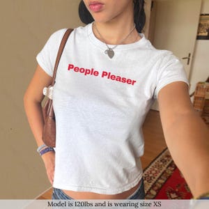 Könnte beinhalten: Weißes, bauchfreies T-Shirt mit dem Text "People Pleaser" in roten Buchstaben.