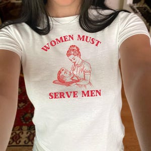 Pode incluir: Camiseta branca com texto vermelho que diz "Women Must Serve Men". O texto é acompanhado por uma ilustração vintage de uma mulher servindo um homem em uma bandeja.