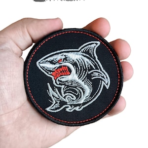 May include: Round black embroidered patch featuring a shark design with white and red accents. The shark has detailed stitching and is set against a black background with a red border. Text at the top reads "DIGITAL EMBROIDERY DESIGN".