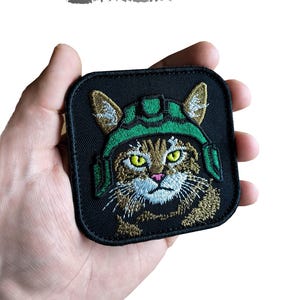 Puede incluir: Parche cuadrado negro con un gato bordado con un casco verde. El gato tiene ojos amarillos y pelaje marrón. El parche tiene las palabras "DIGITAL EMBROIDERY DESIGN" y "Instant Download".