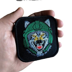 May include: Black square patch with an embroidered wolf wearing a green helmet. The wolf has blue and gray fur, yellow eyes, and a red mouth. The patch has the words "DIGITAL EMBROIDERY DESIGN" and "Instant Download" printed on it.