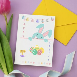 May include: An Easter greeting card with a blue bunny illustration and the words "Happy Easter." The card is accompanied by a yellow envelope, tulips, and a light blue ribbon, all set against a purple background.