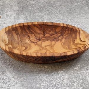 Könnte beinhalten: Ovale Schale aus Olivenholz mit natürlicher Maserung. Die Schale ist hellbraun mit dunkleren Streifen und steht auf einer grauen Oberfläche. Ein dekoratives Element für das Zuhause.