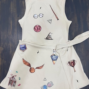 May include: Cream-colored sleeveless dress featuring various Harry Potter-themed graphics. The dress includes images of glasses, a wand, a sorting hat, a snitch, and other iconic symbols from the series. A wrap-around skirt is attached.