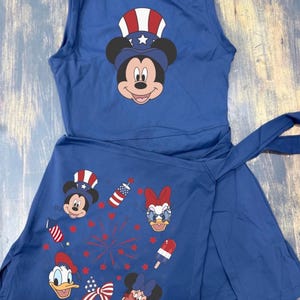May include: A navy blue sleeveless dress featuring Mickey Mouse in a patriotic hat on the bodice. The skirt has a wrap design with images of Mickey, Minnie, Donald, and Daisy Duck, along with fireworks and a popsicle. The dress is perfect for a themed celebration.