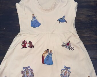 Puede incluir: Vestido sin mangas, blanco roto, con cuello redondo y gráficos temáticos de Cenicienta. El vestido tiene una falda acampanada y presenta ilustraciones de Cenicienta, el zapato de cristal y otros personajes en azul y rojo.