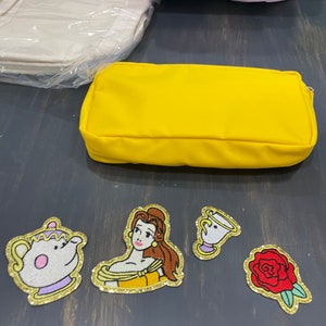 Puede incluir: Estuche de lápices de tela amarillo con parches bordados de Bella de La Bella y la Bestia, una taza de té y una rosa roja.