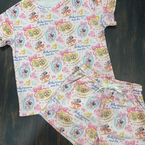 May include: A pink two-piece pajama set featuring a repeating pattern of cartoon characters, teacups, bows, and the text "Girls just wanna have fun." The set includes a short-sleeved top and matching shorts with a drawstring waist.