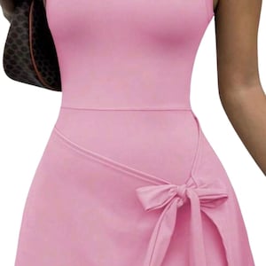 May include: Pale pink sleeveless dress with a white cartoon mouse outline on the chest. The dress has a wrap-style skirt with a bow detail. A brown handbag is visible in the background.