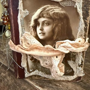 Könnte beinhalten: Vintage-Journal mit einem Sepia-Porträt einer jungen Person. Das Porträt ist mit zarter Spitze und einem rosafarbenen Band gerahmt. Der Einband des Journals ist weinrot, mit einem Perlenanhänger.