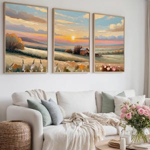 Puede incluir: Tres pinturas de paisajes enmarcadas representan una puesta de sol sobre campos y una pequeña casa. La obra de arte presenta colores cálidos como el naranja, el amarillo y el rosa en el cielo, con campos verdes y marrones debajo. Las pinturas están sobre un sofá blanco.