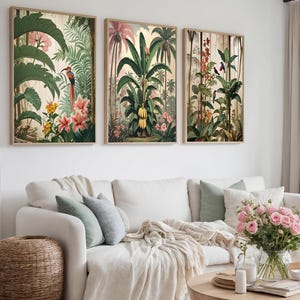 Może przedstawiać: Trzy oprawione botaniczne wydruki przedstawiające bujne tropikalne liście, w tym palmy, kwiaty i ptaki. Grafika ma styl vintage z ciepłą paletą kolorów, wyświetlaną nad białą sofą z dekoracyjnymi poduszkami.