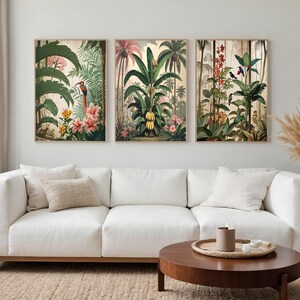 Three framed botanical prints depicting tropical scenes with plants and birds. The artwork features vibrant green foliage, pink and yellow flowers, and various birds. The prints are displayed above a white sofa in a living room.