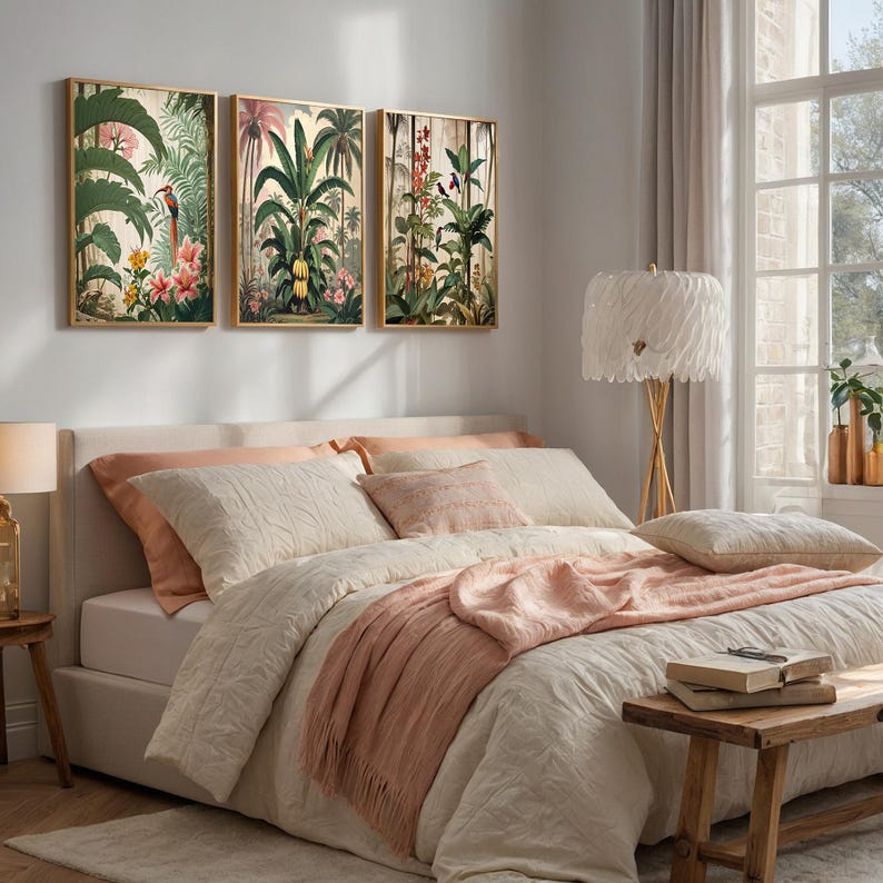 A bedroom with a bed dressed in cream bedding, peach pillows, and a fringed throw. Above the bed, three framed botanical prints with gold frames showcase tropical plants and flowers. A wooden bench holds books.