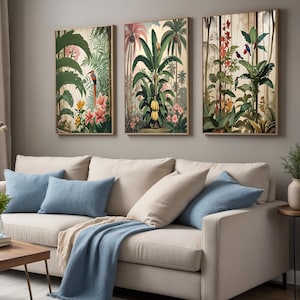 Three framed botanical prints showcasing tropical scenes with green foliage, pink flowers, and birds. The artwork hangs above a beige sofa with blue cushions and a blue throw. The prints are in a living room.