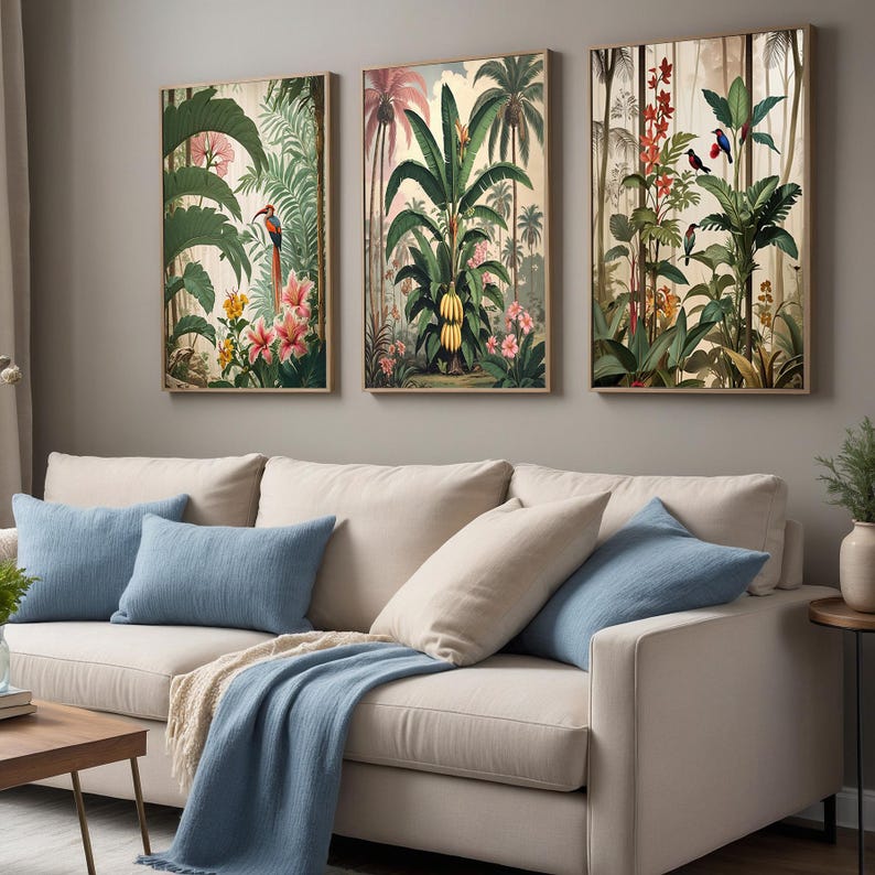 Three framed botanical prints showcasing tropical scenes with green foliage, pink flowers, and birds. The artwork hangs above a beige sofa with blue cushions and a blue throw. The prints are in a living room.